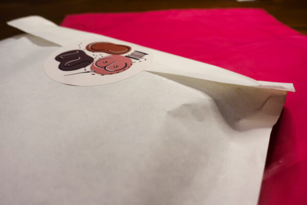 The Jet Set Kit is packaged in a white paper bag, sealed with a sticker featuring the Sew Busty logo of three illustrations of different breasts, a spool of thread, a needle, and stitches, set atop a neon pink mailer.