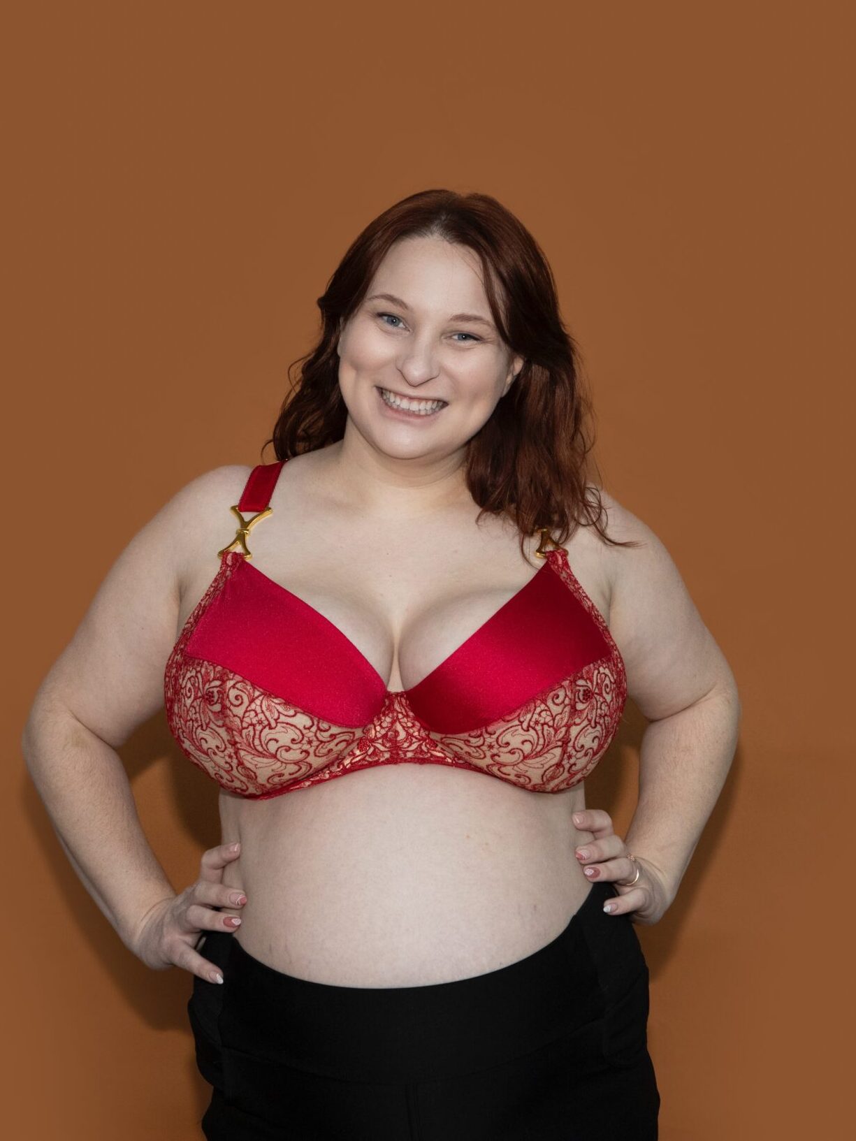 A smiling woman with reddish-brown hair wears a striking red and gold Madalynne Fiona Bra and black pants, standing against a plain brown background with her hands on her hips.