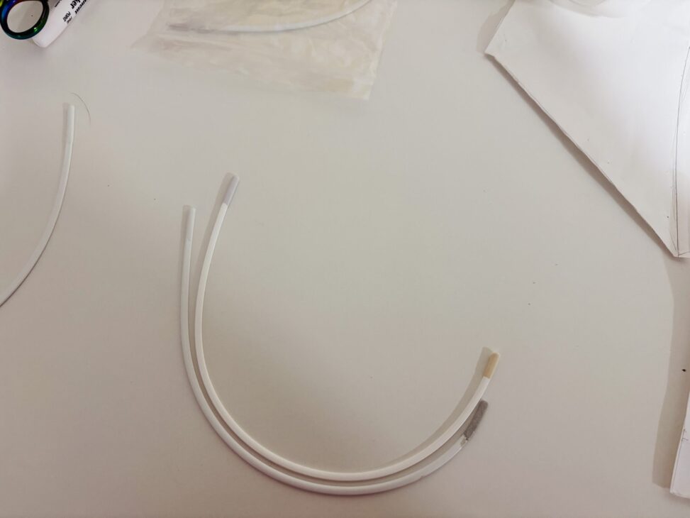 Three white, flexible plastic tubes with different colored ends—one clear, one tan, and one gray—are placed in a loose arc on a white surface, alongside papers and items including a Pattern Review for the Madalynne Fiona Bra.