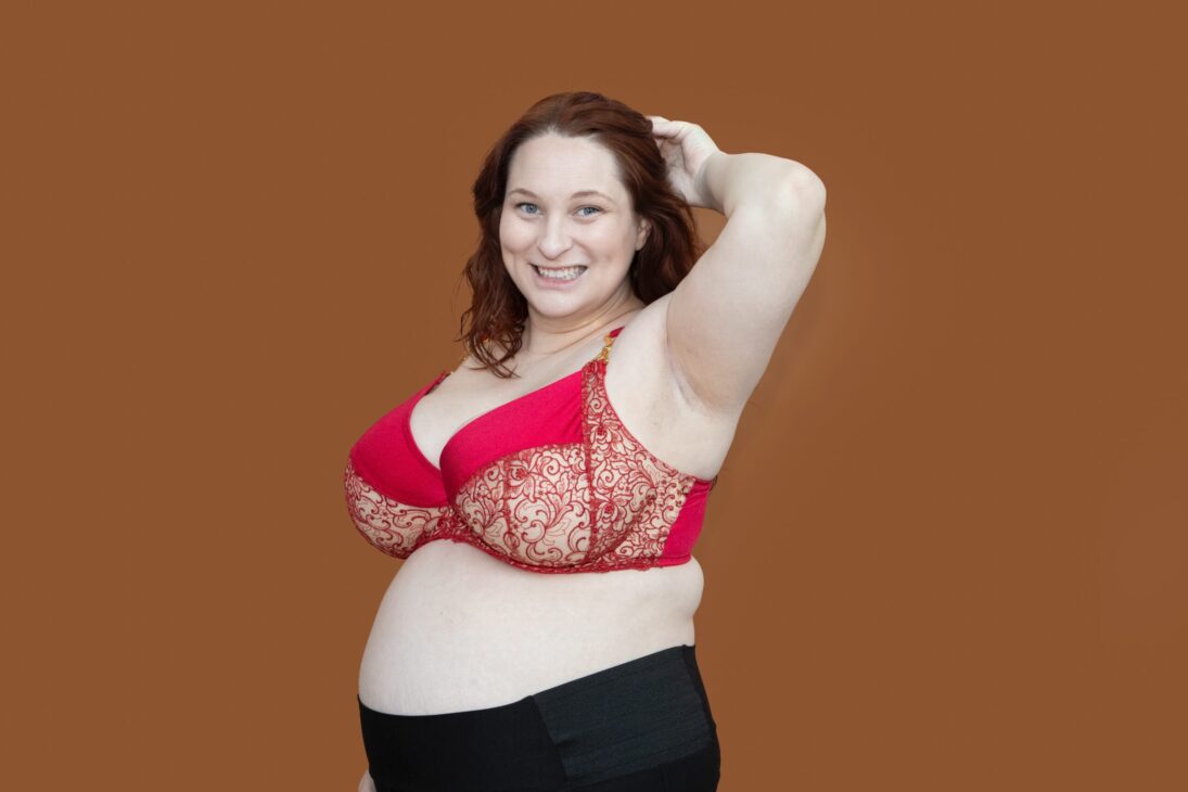 A smiling woman with fair skin and red hair poses confidently with one hand behind her head, modeling the Madalynne Fiona Bra and black bottoms against a plain brown background for a Pattern Review.