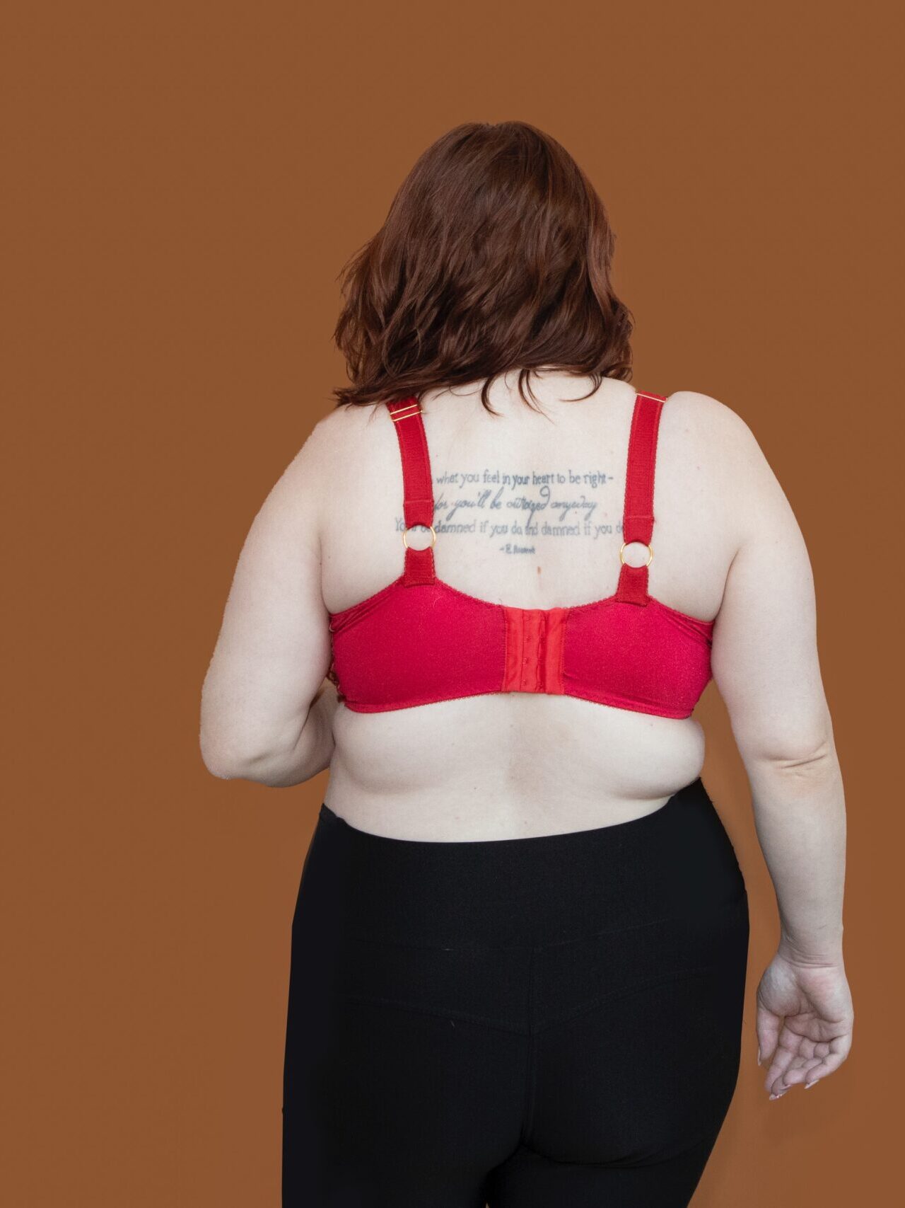 A person with shoulder-length brown hair, wearing a red Fiona Bra and black pants, stands with their back to the camera against a plain brown background. A tattoo with text is visible on their upper back.